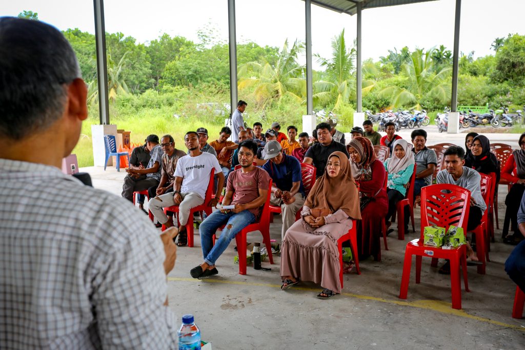 A group of 30 people or so are sitting on red chairs under a large gazebo while participating in a community meeting. A facilitator’s back is visible in the foreground.