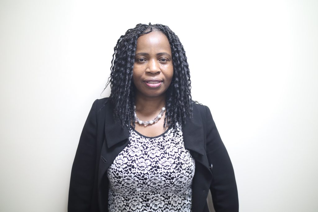 Alt Text: A photo of Dr Adelaide Lusambili standing against a white wall. She is wearing a top with black and white flowers and a black blazer over it.