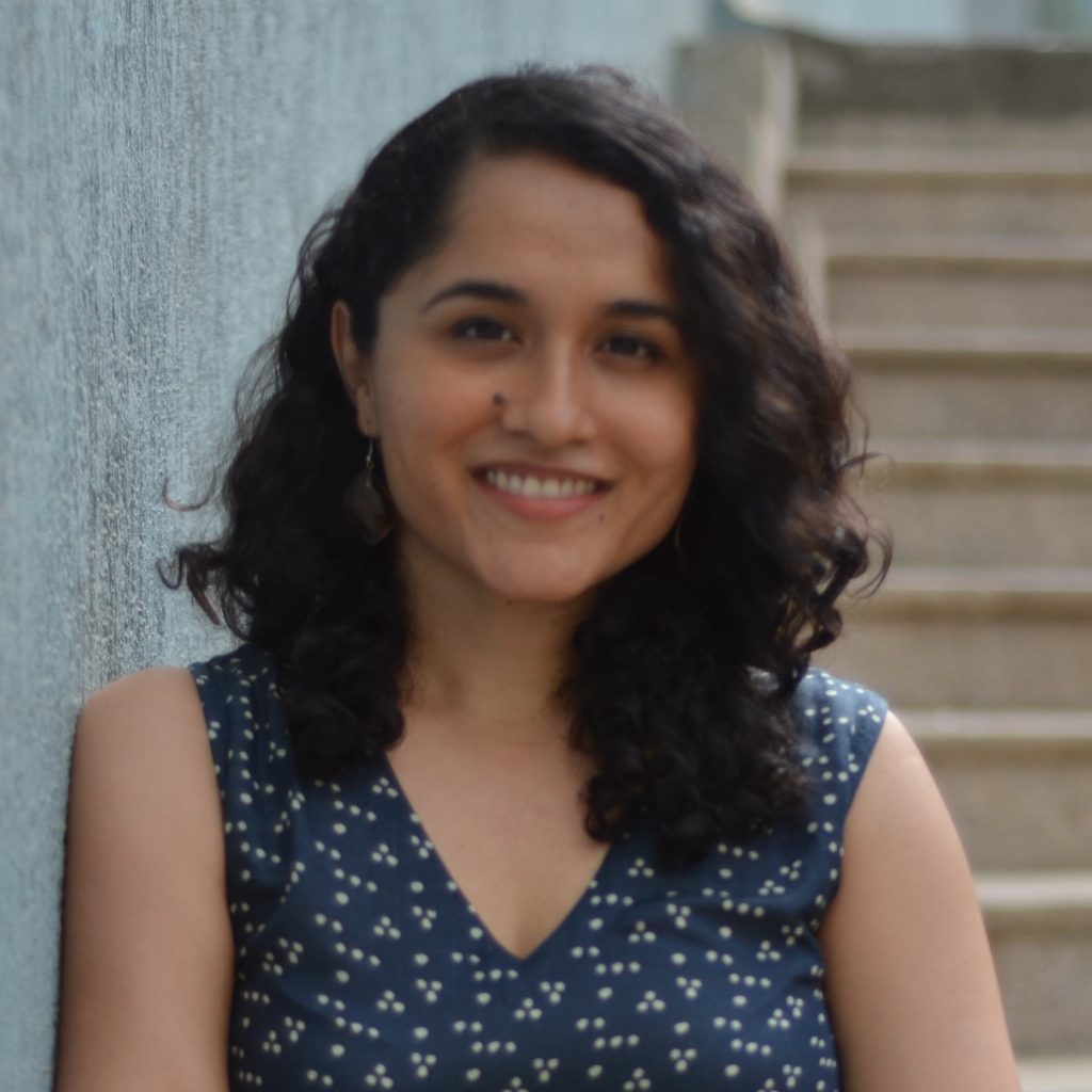 Alt Text: A photo of Amrita Mahale, the Director of Product and Innovation at ARMMAN. She is wearing a blue top with white dotted patterns. In the background is a green wall and a brown staircase.