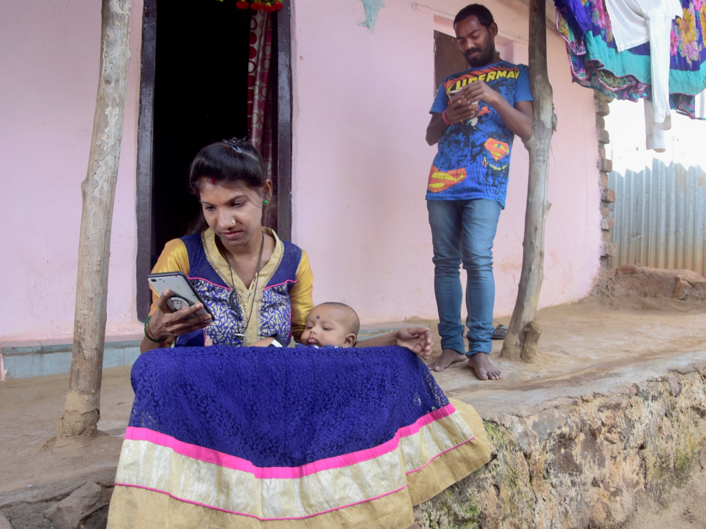 Caption: ARMMAN shares messages regarding maternal and child health over the phone. Photo Credit: ARMMAN Alt Text: A woman in a blue dress scrolls through a phone as she sits outside a house with a baby on her lap. Behind her is a man in a blue shirt and jeans, standing and looking at his phone. The house’s facade is pink and there are clothes drying in the background.